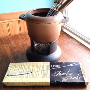 Può includere: Un set per fonduta vintage con una pentola in ceramica marrone, una base nera e sei forchette da fonduta con manici in legno. La scatola delle forchette è etichettata "6 PIECE Fondue Forks".