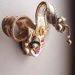 Peut inclure: Un masque de bouffon décoratif avec un visage blanc, des lèvres rouges et des détails d'yeux colorés. Le masque présente une couronne dorée, des cloches et des coiffes à rubans élaborées ornées d'images de cartes de tarot et d'accents turquoise.