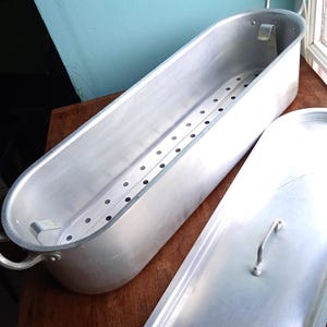 May include: An oval-shaped aluminium fish poacher with a matching lid. The poacher has a perforated interior rack and handles. The lid has a single handle. The poacher is on a wooden surface.