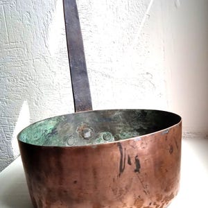 May include: A vintage copper pot with a long iron handle. The pot has a rounded bottom and a wide, shallow opening. The copper has a patina of age and use.