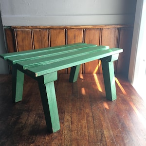 Könnte beinhalten: Ein kleiner, rechteckiger Holzhocker, der in einer soliden grünen Farbe gestrichen ist. Der Hocker verfügt über eine aus drei parallelen Holzlatten bestehende Sitzfläche und wird von vier stabilen, angewinkelten Beinen am Rahmen getragen.
