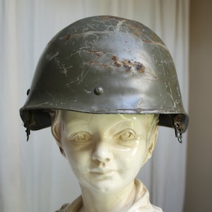 Peut inclure: Un casque militaire vert montrant des signes d'usure. Le casque est en métal et possède une jugulaire.