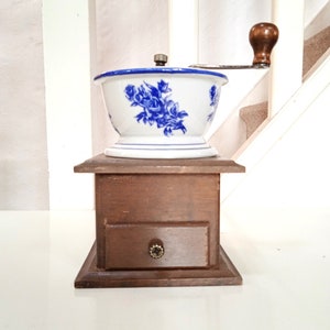 Op de afbeelding: Een vintage houten koffiemolen met een wit keramisch bovendeel met een blauw bloemenmotief. De molen heeft een handmatige slinger en een klein lade.