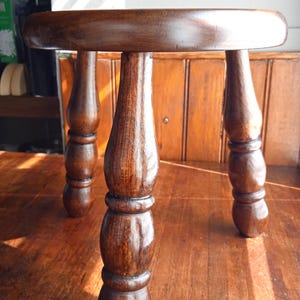 May include: A small, round wooden stool with a dark brown finish. The stool has three turned legs and a circular seat. The legs are intricately carved with a bulbous design. The stool is likely made of hardwood.