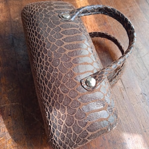 May include: A brown, rectangular case with a snakeskin texture. It features a curved handle and a silver button clasp. The case is designed for glasses or sunglasses. The case is approximately 20 cm long.