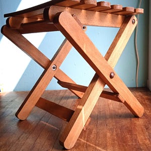 May include: A small, folding wooden stool with a slatted seat. The stool is constructed from light brown wood and features an X-frame design for the legs. The seat is approximately 12 inches long. The stool is designed to be compact and portable.