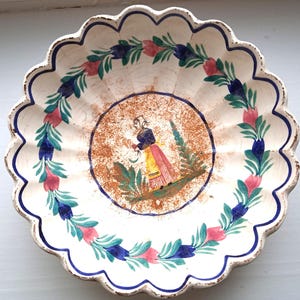 May include: A white ceramic plate with a scalloped edge. The plate is decorated with a blue and green floral design and a central image of a woman in a yellow skirt and blue bodice. The plate has a rustic, vintage look.