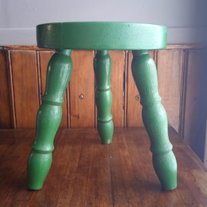 Op de afbeelding: Een kleine, ronde, groene houten kruk met drie gebogen poten. De zitting van de kruk is groen geschilderd en de poten zijn gedraaid en passend geschilderd. De kruk is een vintage stuk, waarschijnlijk gebruikt om op te zitten of als decoratief item.