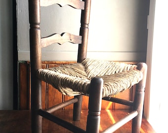 May include: A small wooden chair featuring a dark brown finish, a woven rush seat, and two horizontal back slats with curved edges. The chair has four cylindrical legs connected by straight stretchers and two tall, rounded finial posts.