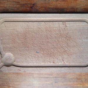 Può includere: Tagliere rettangolare in legno con scanalatura per il succo e manico. La tavola presenta numerosi segni di coltello, a indicare un uso frequente. Il legno ha un tono caldo e naturale, che suggerisce che sia fatto di legno duro.