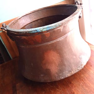 May include: A large, antique copper cauldron with a wide, flared body and a slightly curved rim. The copper has a rich, aged patina with areas of verdigris. Two ornate handles are attached to the rim.