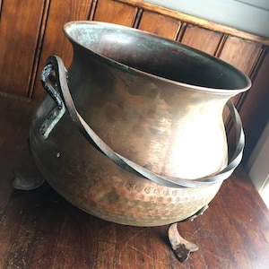 May include: A round, hammered copper cauldron featuring a wide, flared rim with visible green oxidation. The vessel is supported by three curved metal feet and includes a thick, twisted metal handle attached to two side brackets on the body.