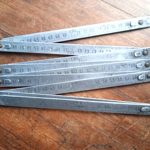Op de afbeelding: Een opengevouwen, zilverkleurige vouwlat met zwarte meetmarkeringen. De metalen lat ligt op een houten oppervlak. De afmetingen zijn in centimeters.