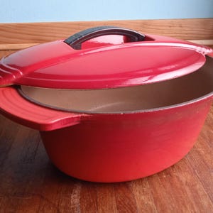 以下が含まれることがあります： 赤色のエナメル鋳鉄製ダッチオーブンと、お揃いの蓋。蓋には黒いハンドルと曲線的なデザインがあります。鍋には両側にハンドルが2つあり、木製の表面に置かれています。