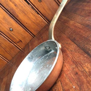 May include: A round copper skillet with a long, curved brass handle. The interior of the pan features a silver-toned metallic lining with visible signs of wear. The exterior sides are polished copper, and the handle is attached with two rivets.
