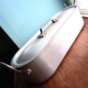 Op de afbeelding: Een grote, ovale aluminium braadpan met twee handvatten aan elke kant en een deksel. De pan is zilverkleurig en heeft een glad oppervlak.