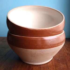 Puede incluir: Dos cuencos de cerámica marrón y blanco apilados uno encima del otro. Los cuencos tienen un borde marrón y un interior blanco.