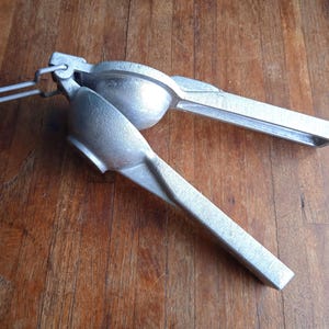 May include: A silver metal citrus juicer. The juicer is open, revealing the two halves connected by a hinge and a handle. It is positioned on a wooden surface.