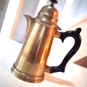 Könnte beinhalten: Eine goldfarbene Kaffeekanne mit schwarzem Griff. Die Kanne hat einen runden Korpus und einen dekorativen Deckel.
