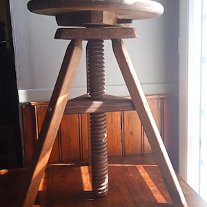 Puede incluir: Taburete de madera ajustable con asiento redondo y columna central roscada. El taburete tiene tres patas anguladas y un acabado marrón oscuro. El asiento tiene unos 30 cm de diámetro.