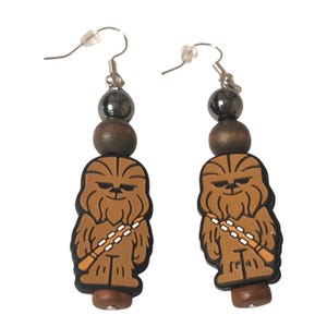 May include: A pair of dangle earrings featuring a brown, tan, and black rubberized Chewbacca character charm. Each earring includes a dark metallic gray bead, a smooth brown wooden bead, and a small brown wooden bead at the bottom base.