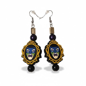 May include: A pair of dangle earrings featuring a black, yellow, and green mask design within a gold-colored frame. The earrings have a black bead at the bottom and a purple bead and gold-colored square above the frame. The earrings hang from silver-colored hooks.