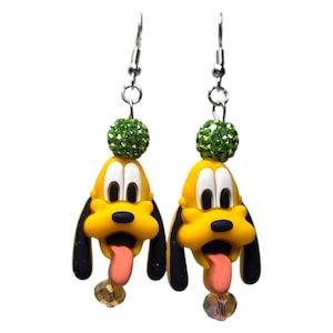 May include: A pair of dangle earrings featuring a yellow cartoon dog head with long black ears, white eyes, and a pink tongue. Above the dog head is a round green glitter bead, and below the tongue is a small, faceted, translucent amber bead.