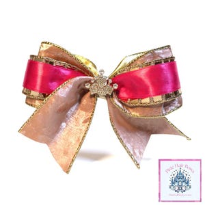 May include: A large, decorative hair bow with layers of gold, pink, and champagne-colored ribbons. A small, jeweled crown is centered on the bow. The bow is on a white background.