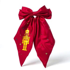 May include: A large, red satin bow with a gold nutcracker soldier design and scattered gold and jewel-toned embellishments. The bow is a festive accessory, perfect for holiday or special occasion wear.