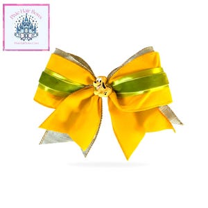 May include: A large yellow hair bow with a green ribbon stripe and silver trim. The bow features a small Pluto character embellishment in the center. The bow is likely for a child.