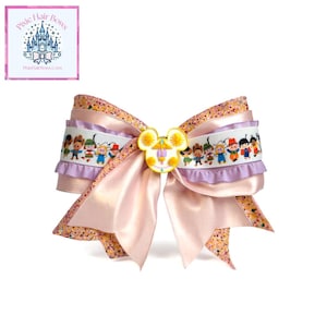 May include: A large, layered hair bow with a light pink satin base and a decorative Mickey Mouse-shaped embellishment. The bow features a white ribbon with a cartoon illustration of characters, and is trimmed with glittery fabric.