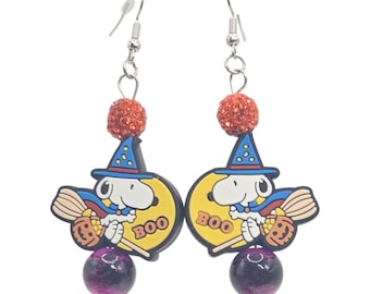 May include: A pair of dangle earrings featuring a white cartoon dog wearing a blue witch hat, riding a broomstick in front of a yellow moon with the text BOO. Each earring includes an orange glitter bead and a purple marbled bead on a silver hook.