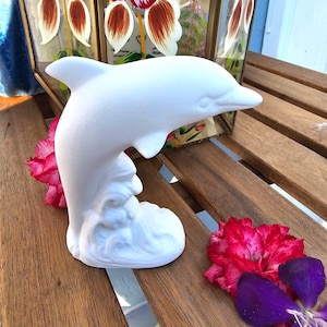 May include: A matte white ceramic dolphin figurine depicted in a leaping motion, emerging from a sculpted wave base. The dolphin has a smooth, unpainted surface and is positioned in a side profile view, showcasing its dorsal fin and snout.
