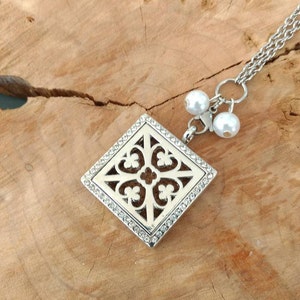 May include: A silver necklace with a square pendant featuring a decorative design and rhinestones. The pendant is attached to a silver chain with two white pearl accents.