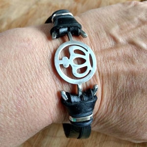 May include: A black leather bracelet with a silver metal crown charm. The bracelet is adjustable and has a simple design.