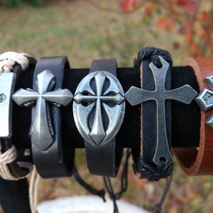 May include: Five leather bracelets with silver cross charms. The bracelets are in various styles, including braided leather, plain leather, and leather with a woven cord. The crosses are all different designs, including a simple cross, a cross with a circle around it, and a cross with a fleur-de-lis design.