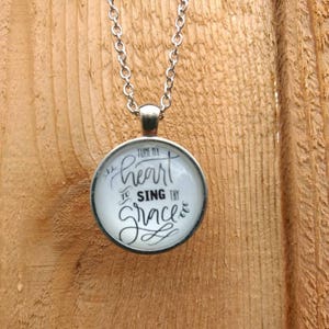 May include: A silver necklace with a round pendant featuring the text "Tune my heart to sing thy grace" in black calligraphy.