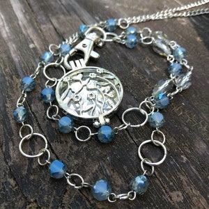 May include: A silver-toned locket necklace with a round, openwork pendant. The pendant is connected to a chain and a beaded strand with blue-gray faceted beads. The necklace has a lobster clasp and is displayed on a wooden surface.