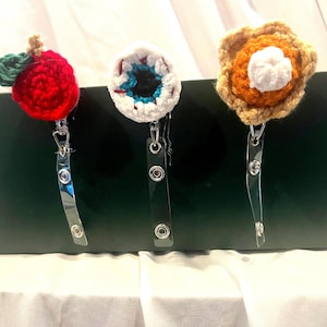 May include: Three badge reels featuring crocheted toppers. The left topper is a red apple with a green leaf. The center topper is a white, blue, and black eyeball. The right topper is a tan and orange pumpkin pie slice with a white dollop of cream.
