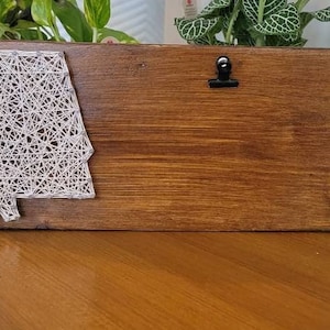 May include: A wooden picture frame with a white string art design of the state of Alabama. The frame has a clip on the top for hanging a photo or note.