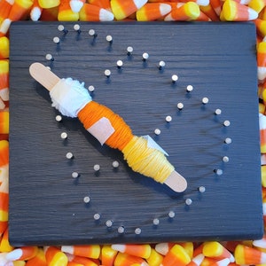 May include: A black wooden board with a heart shape outlined with silver tacks. A multi-colored yarn is wrapped around the tacks, creating a string art design.
