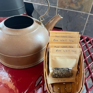 May include: A small wooden basket filled with four brown paper packets of Bee Well Tea. The packets are labeled with the text "Bee Well Tea". The basket is sitting on a red surface next to a copper kettle.