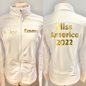 May include: White zippered jacket with gold lettering. The front of the jacket has a crown design and the name "Emma". The back of the jacket says "Miss America 2022".