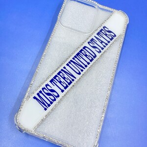 May include: A clear phone case with a white sash that says "Miss Teen United States" in blue lettering. The case is decorated with rhinestones around the edges.