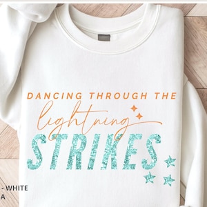 May include: A white crewneck sweatshirt featuring the orange text DANCING THROUGH THE and the word lightning in a cursive orange script. Below, the word STRIKES is printed in a large, glittery teal font, accompanied by small teal star icons.