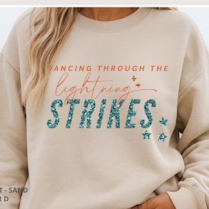May include: A sand-colored crewneck sweatshirt featuring the text DANCING THROUGH THE lightning STRIKES in orange and teal glittery font. The design includes small orange and teal star accents. The top text reads MADE TO ORDER &bull; CHOOSE YOUR STYLE, COLOR & TEXT.