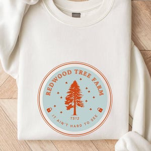 Pode incluir: Um moletom branco com um gr&aacute;fico circular. O gr&aacute;fico diz "REDWOOD TREE FARM" em vermelho, com uma ilustra&ccedil;&atilde;o de &aacute;rvore vermelha, estrelas e a frase "IT AIN'T HARD TO SEE". O moletom tem gola redonda e mangas compridas.