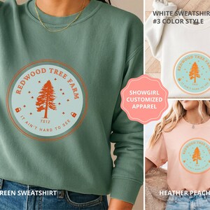 Pode incluir: Uma camisola verde claro com um gr&aacute;fico circular que diz "Redwood Tree Farm" com uma ilustra&ccedil;&atilde;o de &aacute;rvore. O gr&aacute;fico &eacute; em laranja e azul claro. O texto "It ain't hard to see" tamb&eacute;m est&aacute; inclu&iacute;do. Camisas adicionais em branco e p&ecirc;ssego tamb&eacute;m s&atilde;o mostradas.