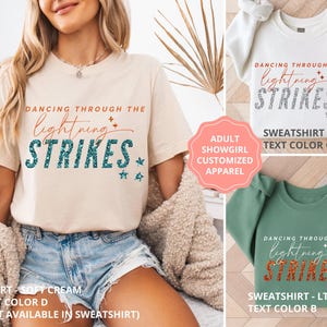 May include: A soft cream t-shirt, a white sweatshirt, and a light green sweatshirt, each featuring the text DANCING THROUGH THE lightning STRIKES with star accents. The t-shirt has teal text, the white sweatshirt has grey text, and the green sweatshirt has orange text.