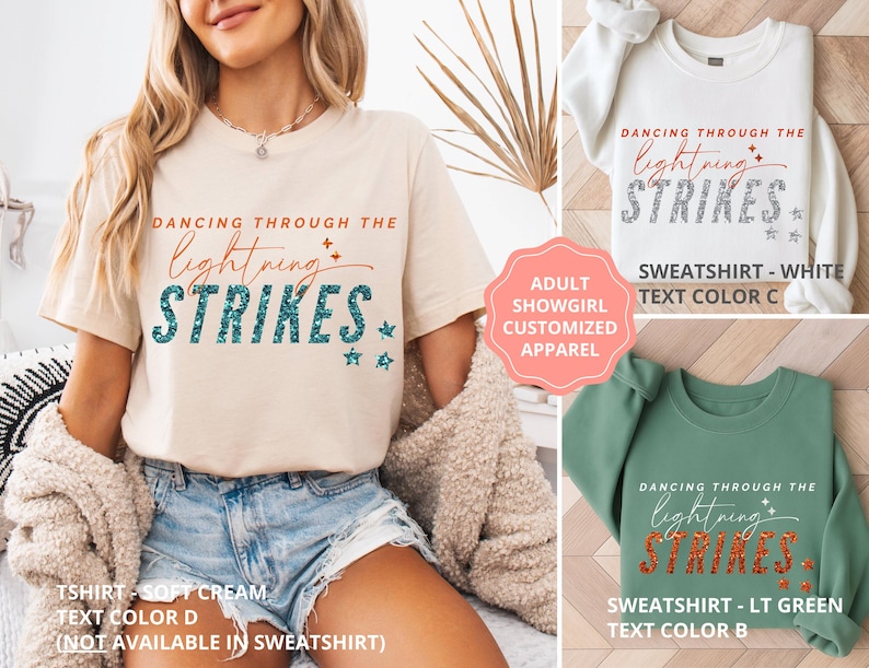May include: A soft cream t-shirt, a white sweatshirt, and a light green sweatshirt, each featuring the text DANCING THROUGH THE lightning STRIKES with star accents. The t-shirt has teal text, the white sweatshirt has grey text, and the green sweatshirt has orange text.